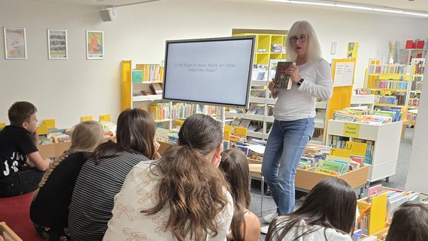 Ein Frau hält ein Buch in der Hand und spricht zu Kindern, die vor ihr sitzen. Neben der Frau steht ein elektronisches Whiteboard. Die Kinder sitzen auf rot bezogenen Treppenstufen mit dem Rücken zum Fotografen. Im Hintergrund sieht man Regale mit Büchern darin. Auf dem Whiteboard steht der Satz "Es fiel Regen in der Nacht, ein feiner, wispernder Regen.". Dies ist der erste Satz aus dem Buch, dass die Frau in der Hand hält: "Tintenherz" von Cornelia Funke.