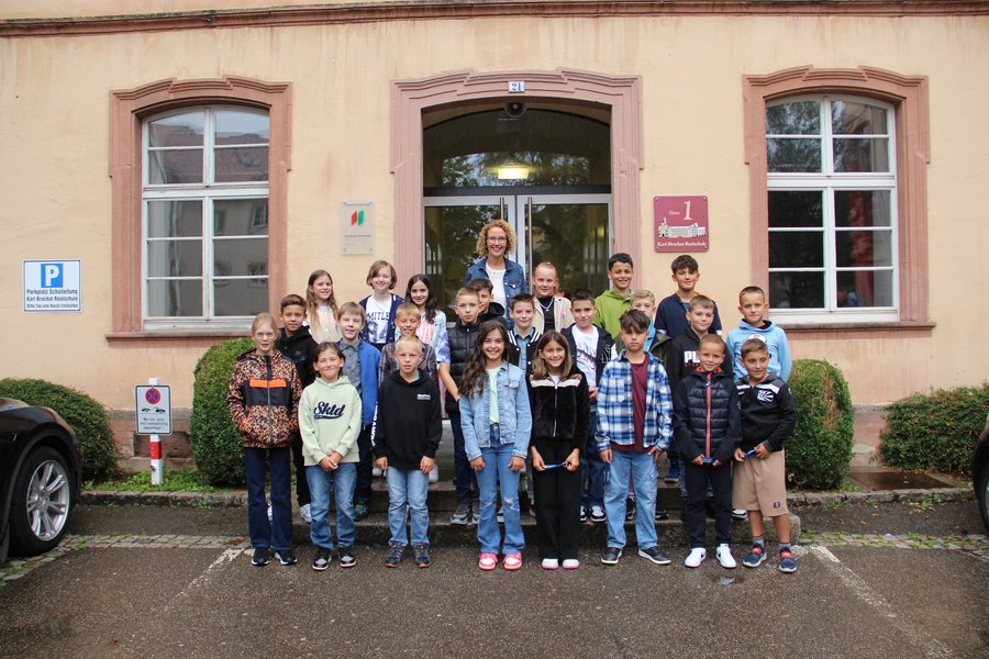 Die Klasse 5c mit ihrer Klassenlehrerin Frau Hildebrand Die Schulkinder stehen in mehreren Reihen auf Treppenstufen vor dem Schuleingang und lächeln in die Kamera.