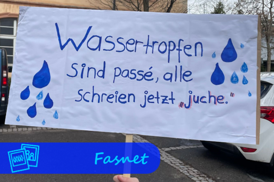 Ein Plakat wird hochgehalten. Auf dieses sind Wassertropfen gemalt und der Mottospruch: "Wassertropfen sind passé, alle schreien jetzt: Juchhe!"